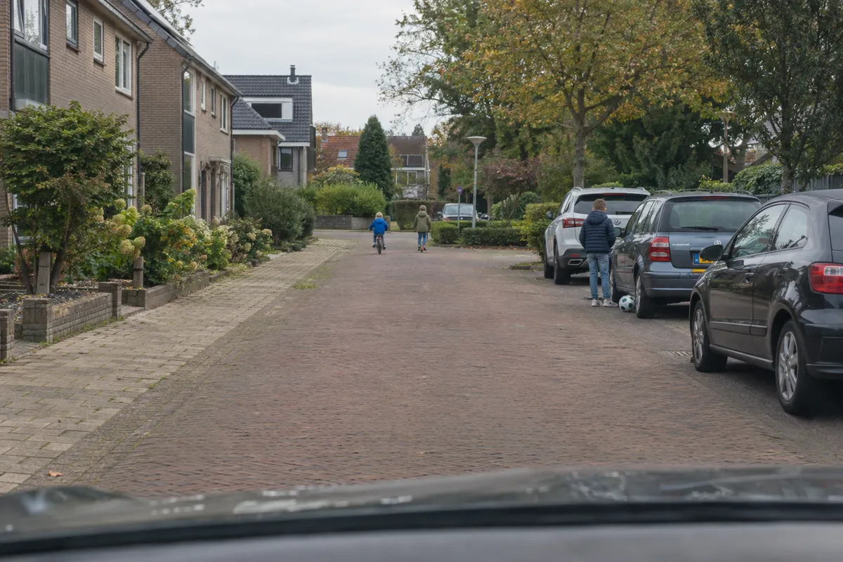 Zicht vanuit de auto in een erf: auto's geparkeerd rechts, een kind op de rug bij een auto, verderop spelende kinderen en een bal op de straat.