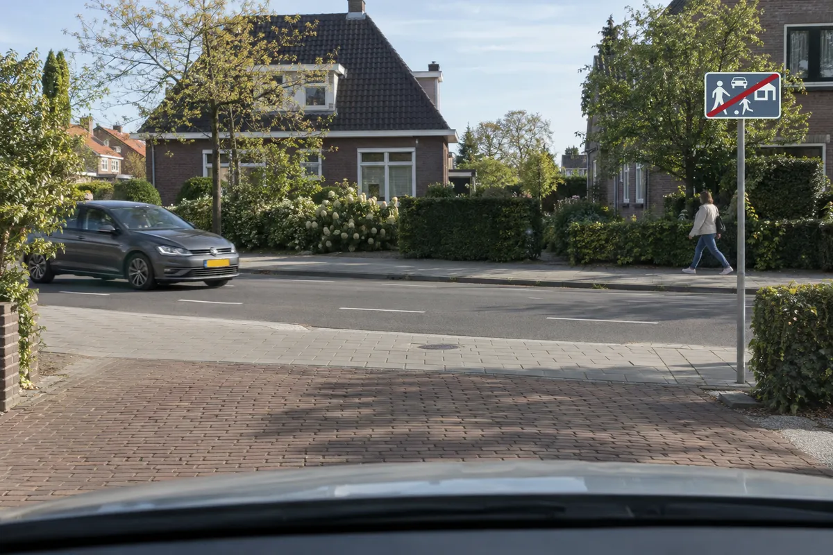Zicht vanuit de auto, nog binnen het erf bij een uitrit. Een voetganger nadert op de stoep en een auto nadert op de weg. Het bord Einde erf staat rechts.