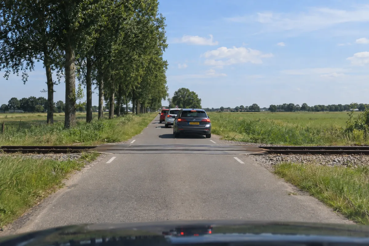Zicht vanuit de auto op een overweg met een spoor dwars over de weg, met vlak achter de rails een stilstaande rij auto's