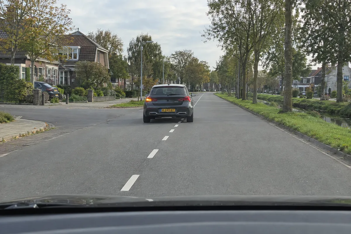 Zicht vanuit de auto op een weg in de bebouwde kom: de auto voor je rijdt links op de weg, zijn linker richtingaanwijzer knippert en rechts van hem is ruimte