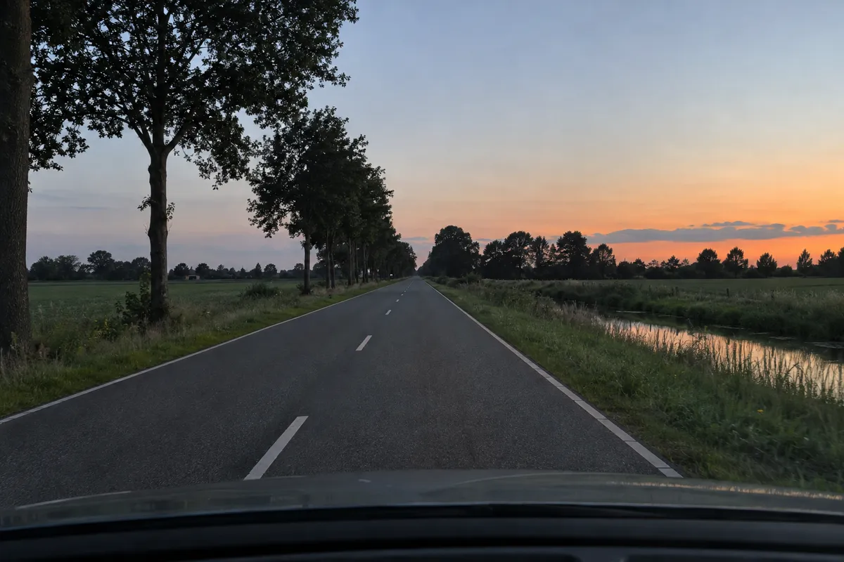 Je staat buiten de bebouwde kom op de rijbaan. De zon is net onder en de omgeving is nog goed te zien.