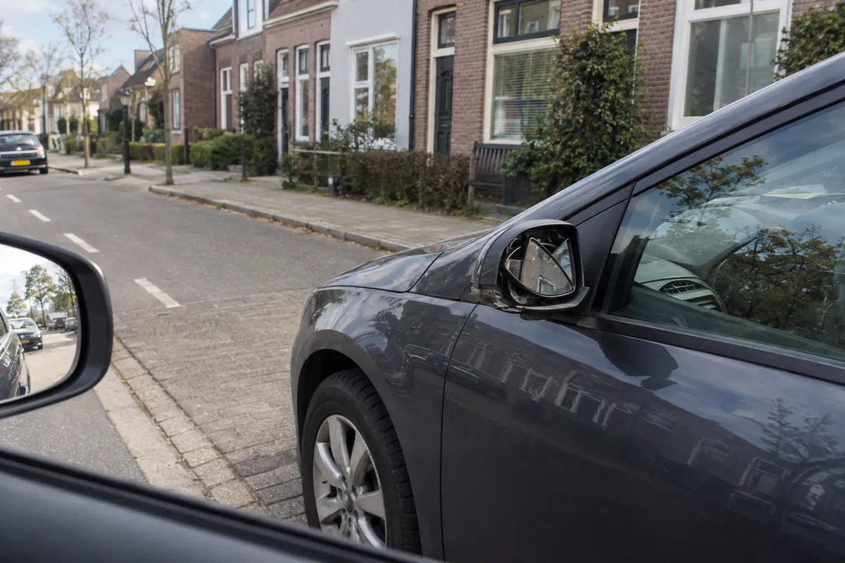 De buitenspiegel van de geparkeerde auto is beschadigd; de eigenaar is niet in beeld.