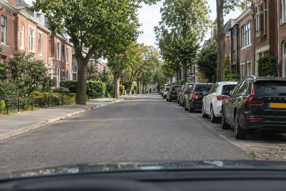 Zicht vanuit de auto op een brede straat: rechts staat een aaneengesloten rij geparkeerde auto’s en jouw auto rijdt op ruime afstand daarvan